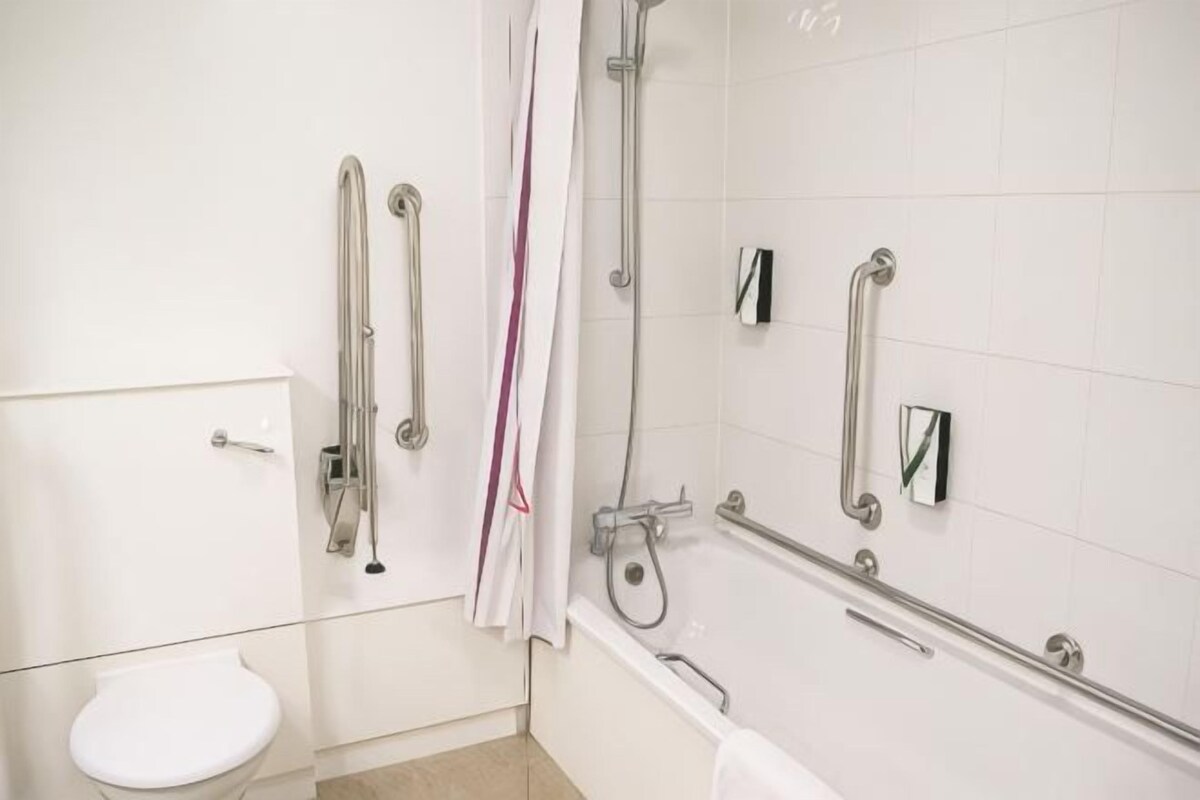 Une salle de bain avec des toilettes blanches, une douche dotée d’une porte en verre et des barres d’appui.
