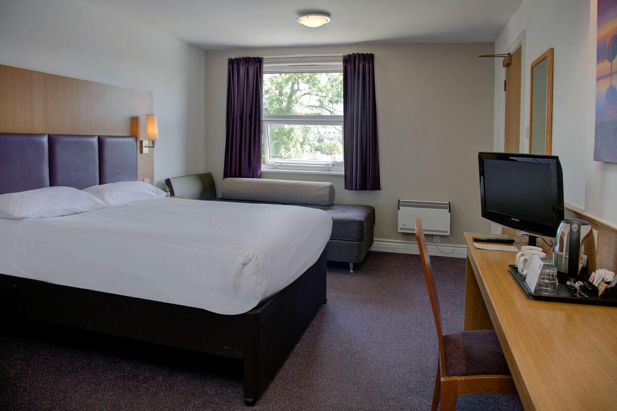 A hotel room with a large bed, a desk with a computer, a chair, and a window with curtains.