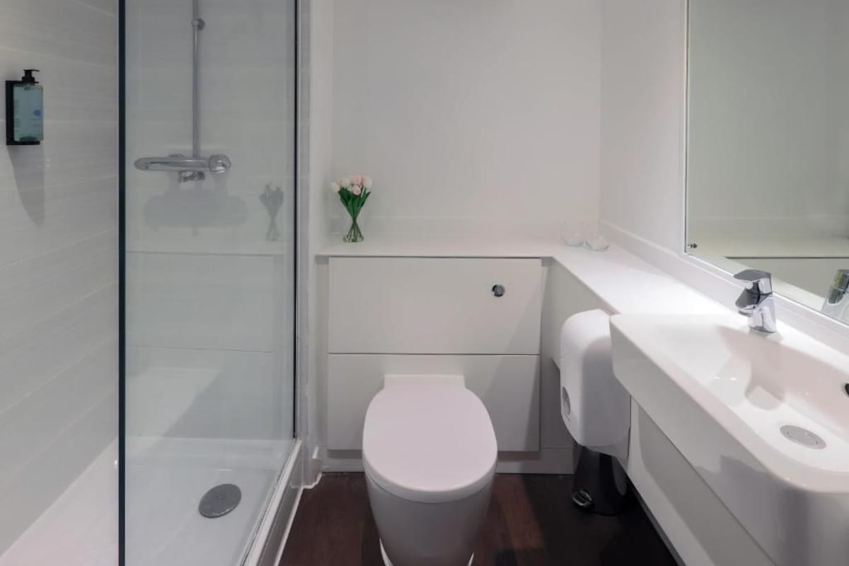 A modern bathroom with a glass shower enclosure, a white toilet, a vanity with a mirror, and a small vase with flowers.