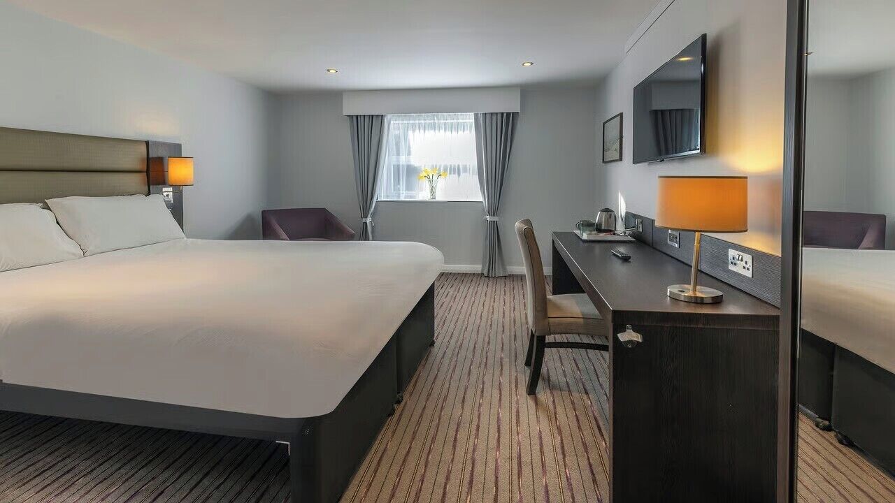 Habitación de hotel con una cama grande, un escritorio con lámpara, una silla, un televisor y una ventana con cortinas.