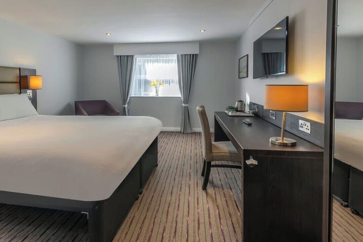 Een hotelkamer met een groot bed, een bureau met lamp, een stoel, een televisie en een raam met gordijnen.