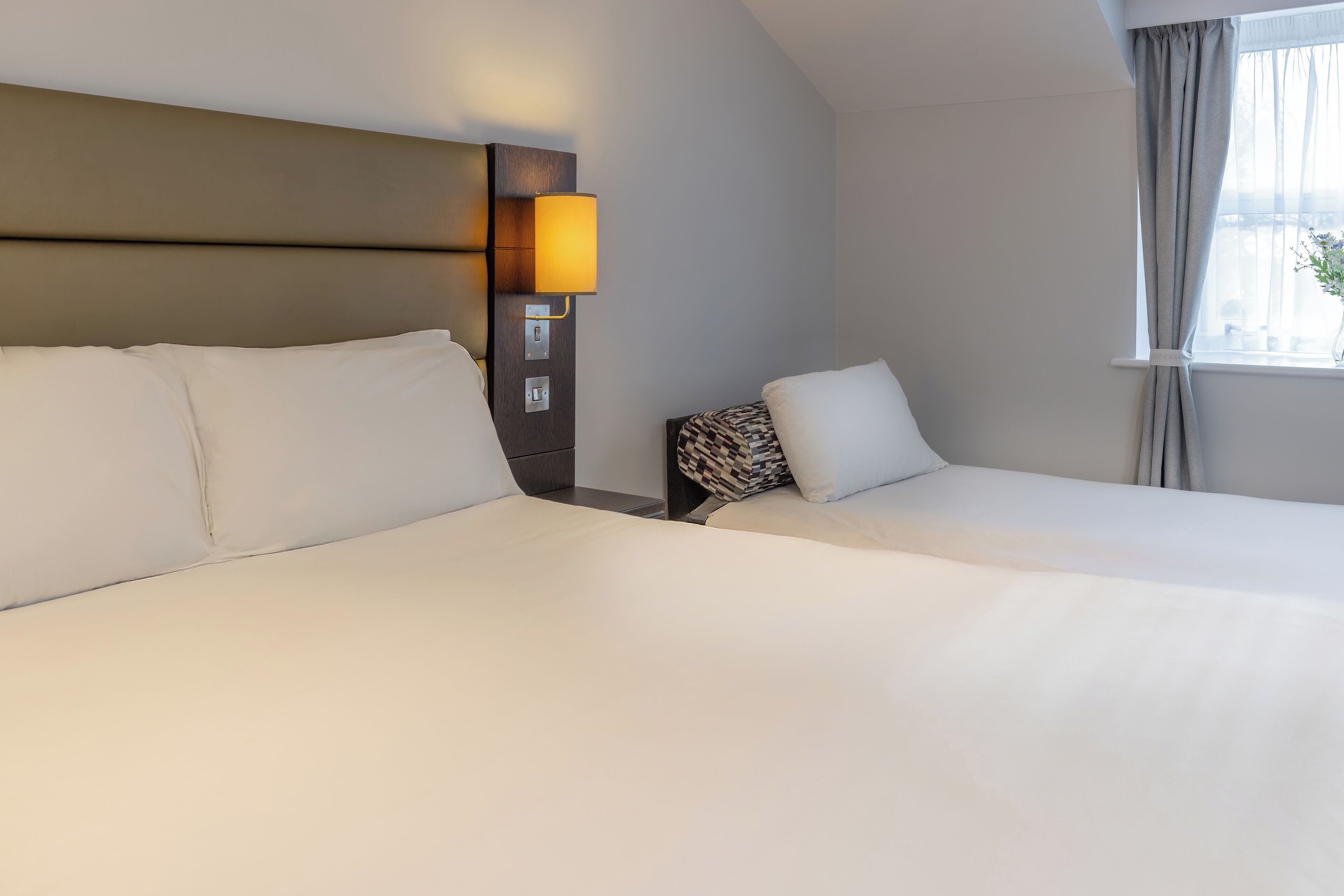 A hotel room with two beds, a headboard with a light, and a window with curtains.