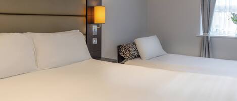 A hotel room with two beds, a headboard with a light, and a window with curtains.