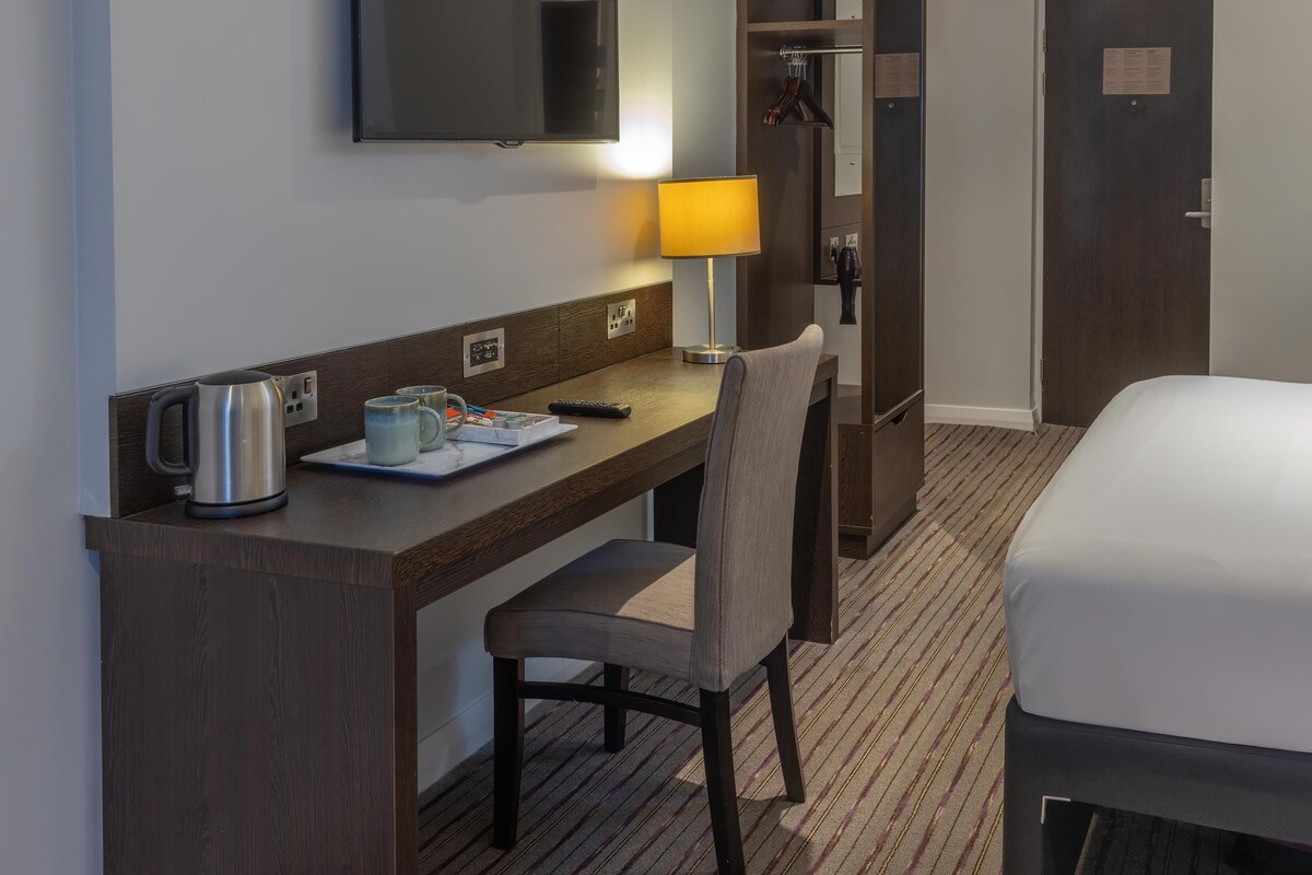 A hotel room with a bed, a desk with a kettle, a chair, a TV, and a lamp.