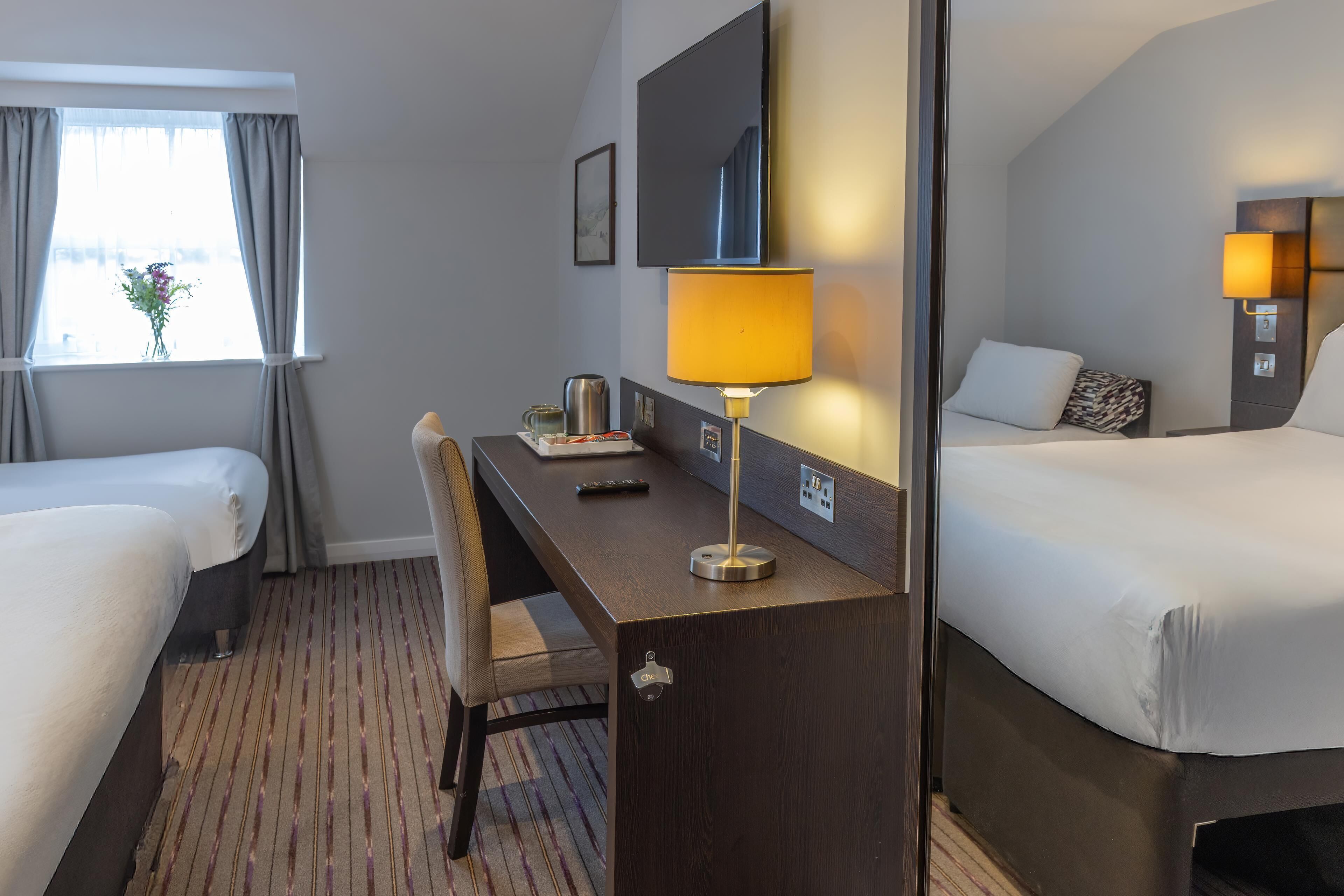 A hotel room with two beds, a desk with a lamp, a chair, a window with curtains, and a wall-mounted TV.