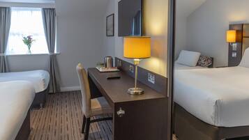 A hotel room with two beds, a desk with a lamp, a chair, a window with curtains, and a wall-mounted TV.