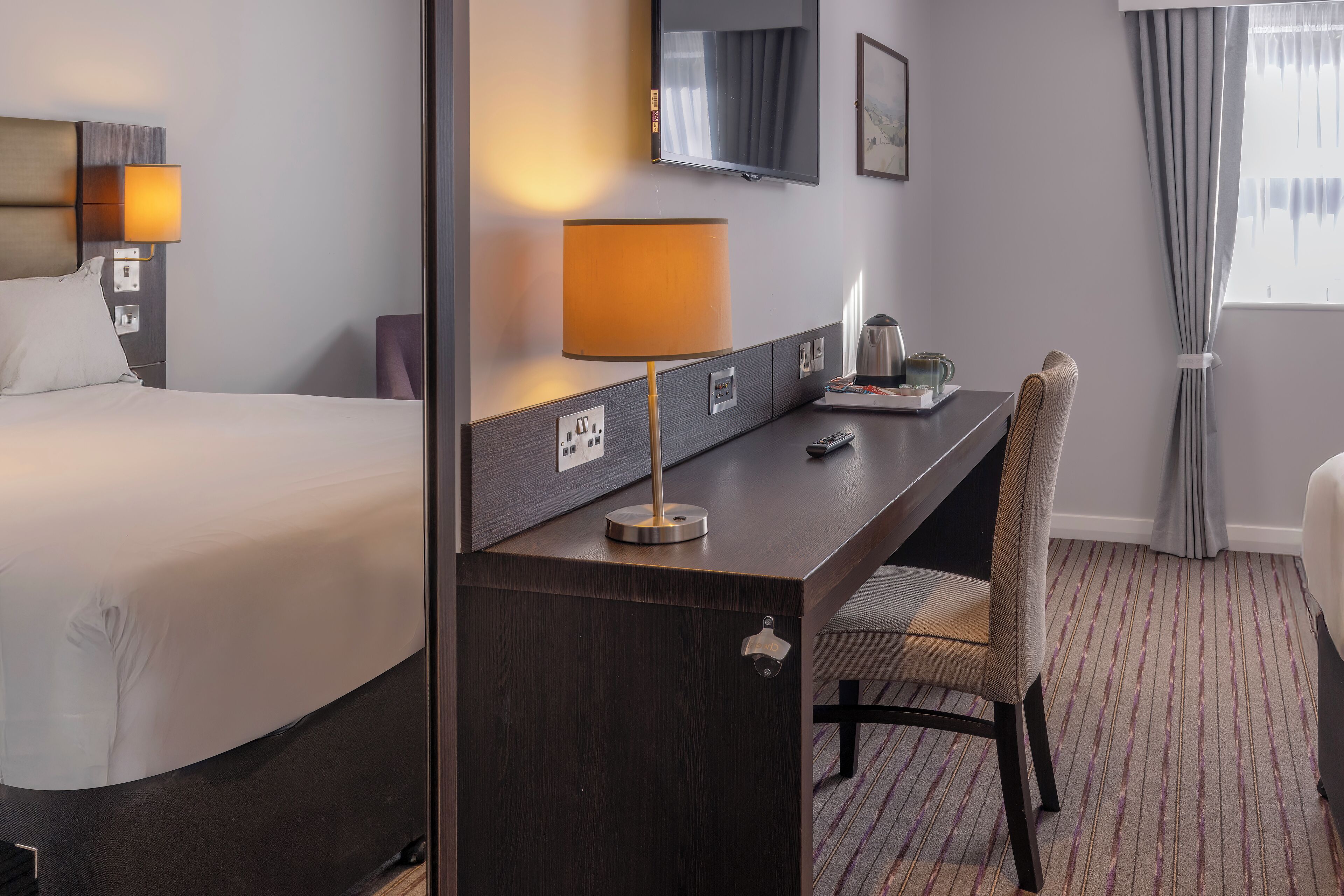 Ein Hotelzimmer mit Bett, Schreibtisch mit Lampe, Stuhl, Fernseher und einem Fenster mit Vorhängen.