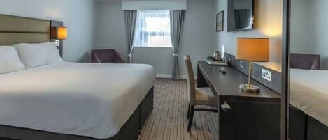 A hotel room with a large bed, a desk, a chair, a desk lamp, a mirror, and a window with curtains.