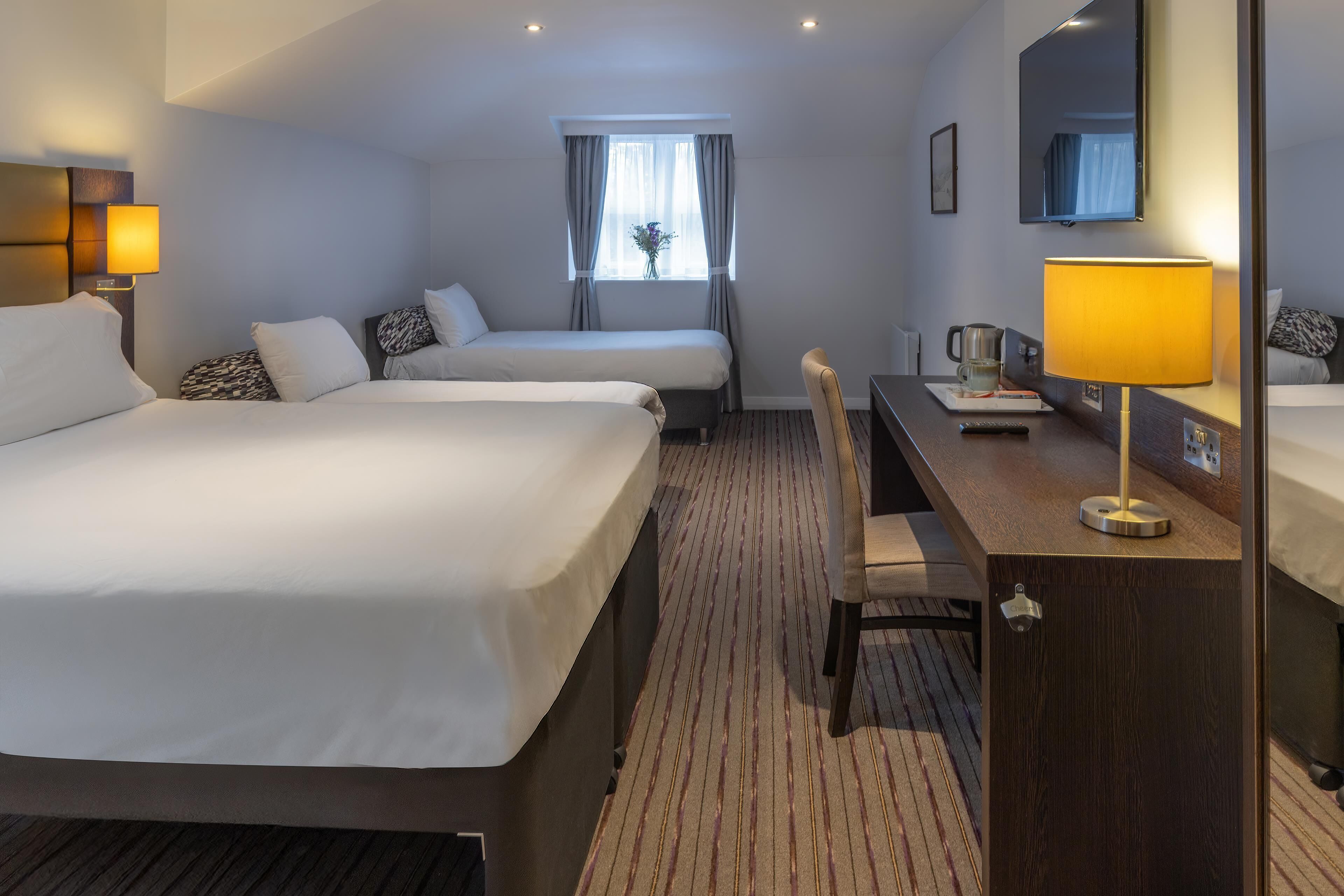 A hotel room with two beds, a desk, a chair, a lamp, a window with curtains, and a mirror.