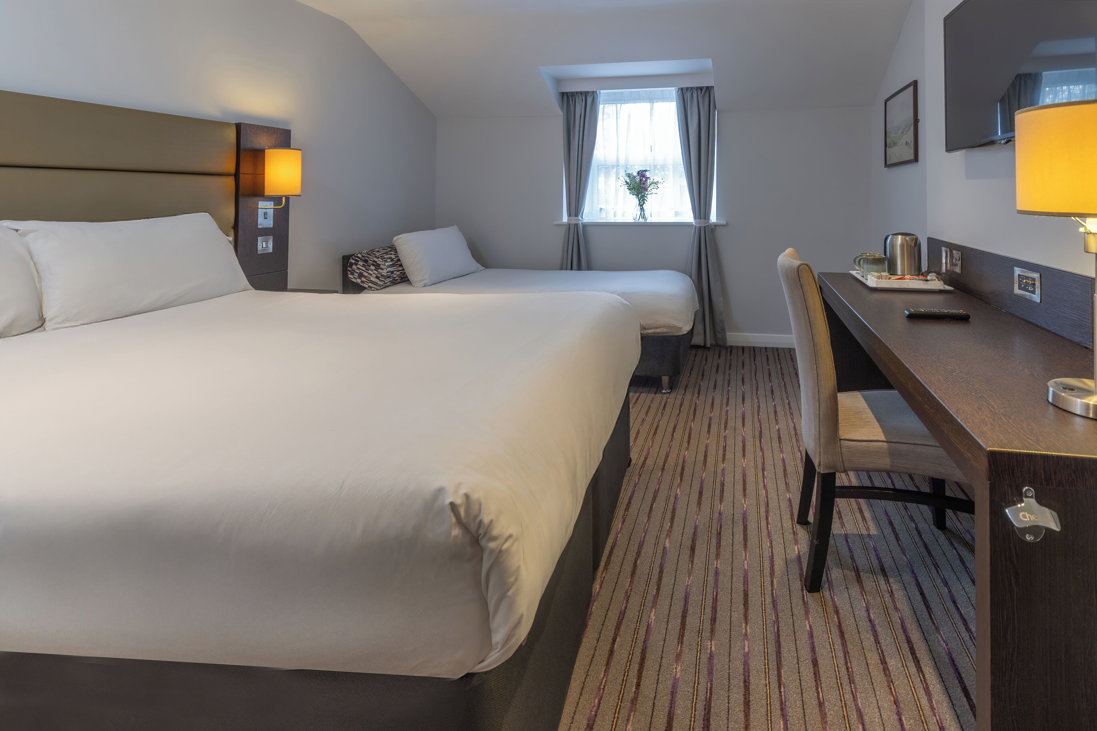 Habitación de hotel con una cama grande, un escritorio con silla, una ventana con cortinas y un televisor de pantalla plana montado en la pared.