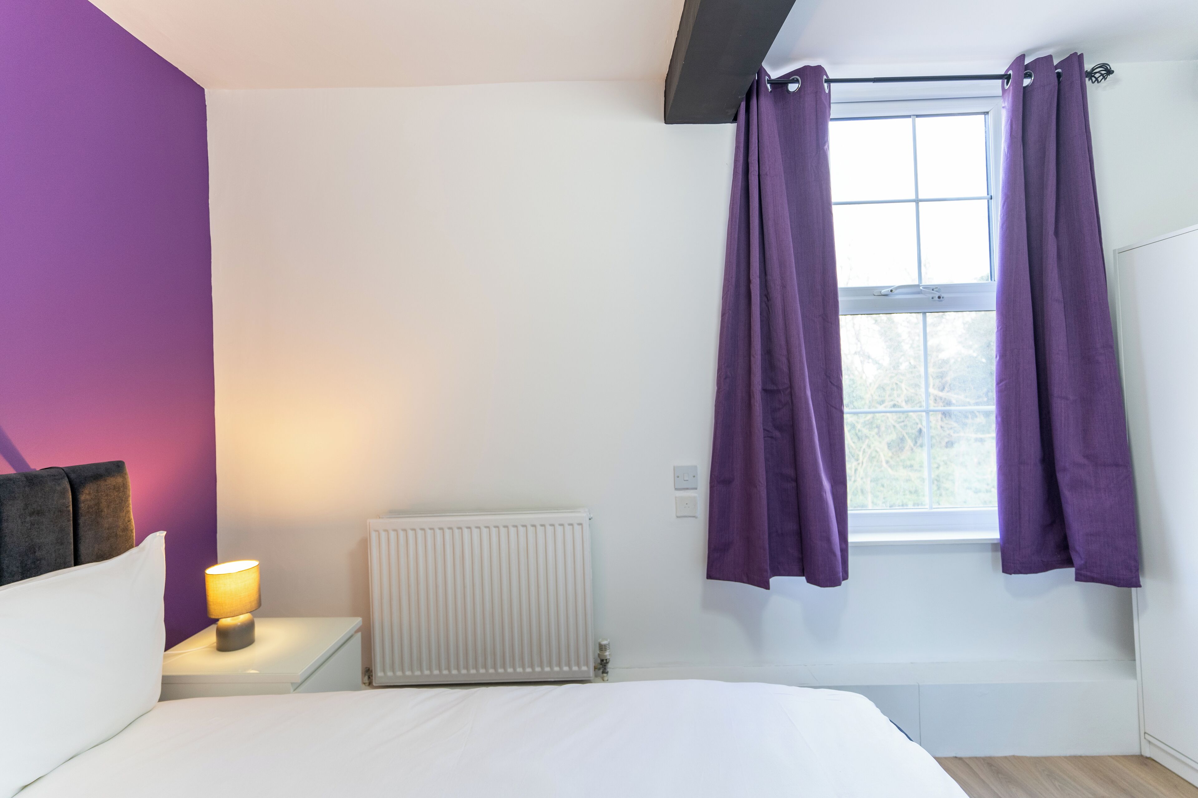 A bedroom with a bed, a nightstand with a lamp, a window with purple curtains, and a radiator.