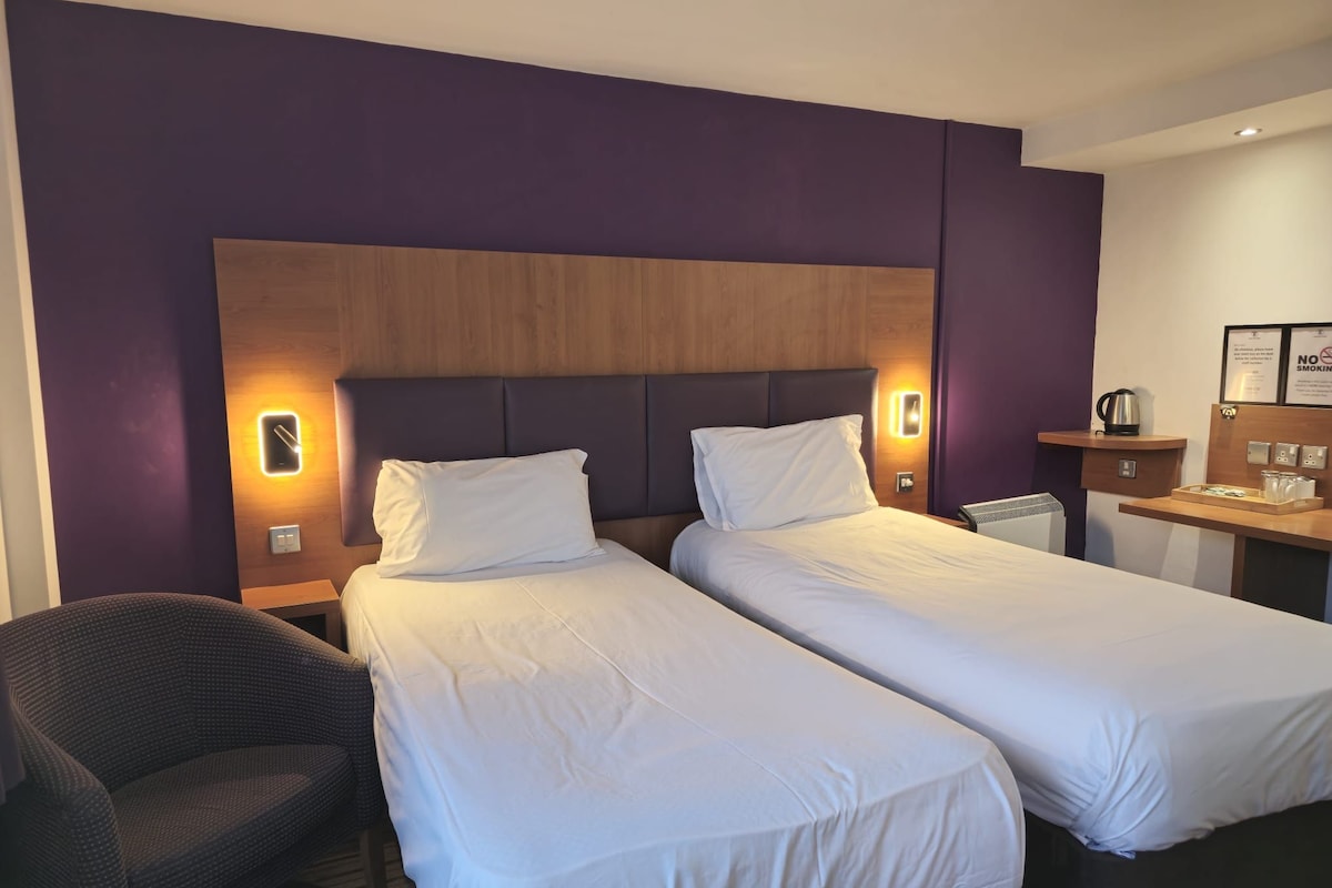 A hotel room with two single beds, a nightstand, a chair, and a small desk with a kettle and a framed sign.