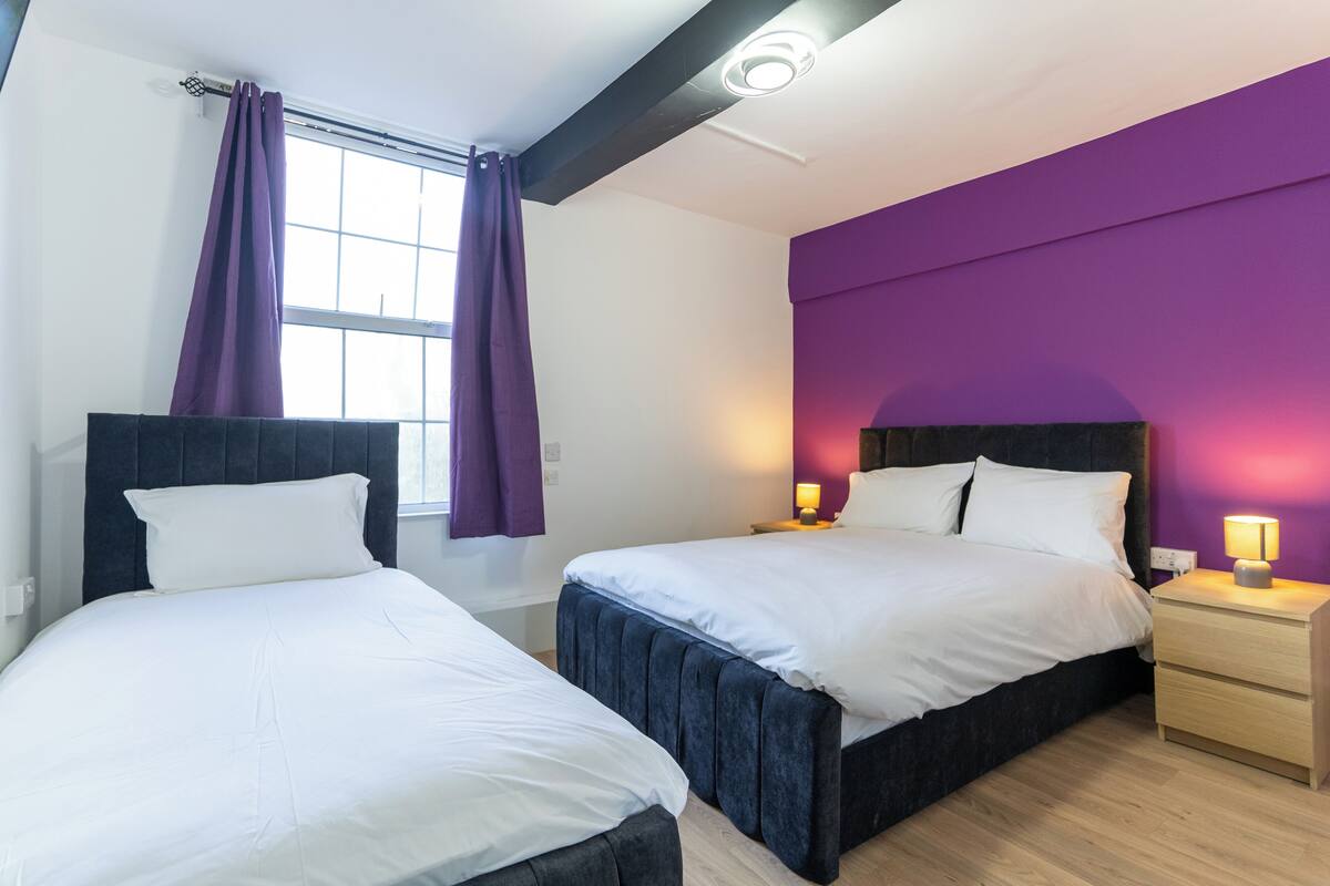 A room with two beds, a window with purple curtains, a purple accent wall, and bedside lamps.
