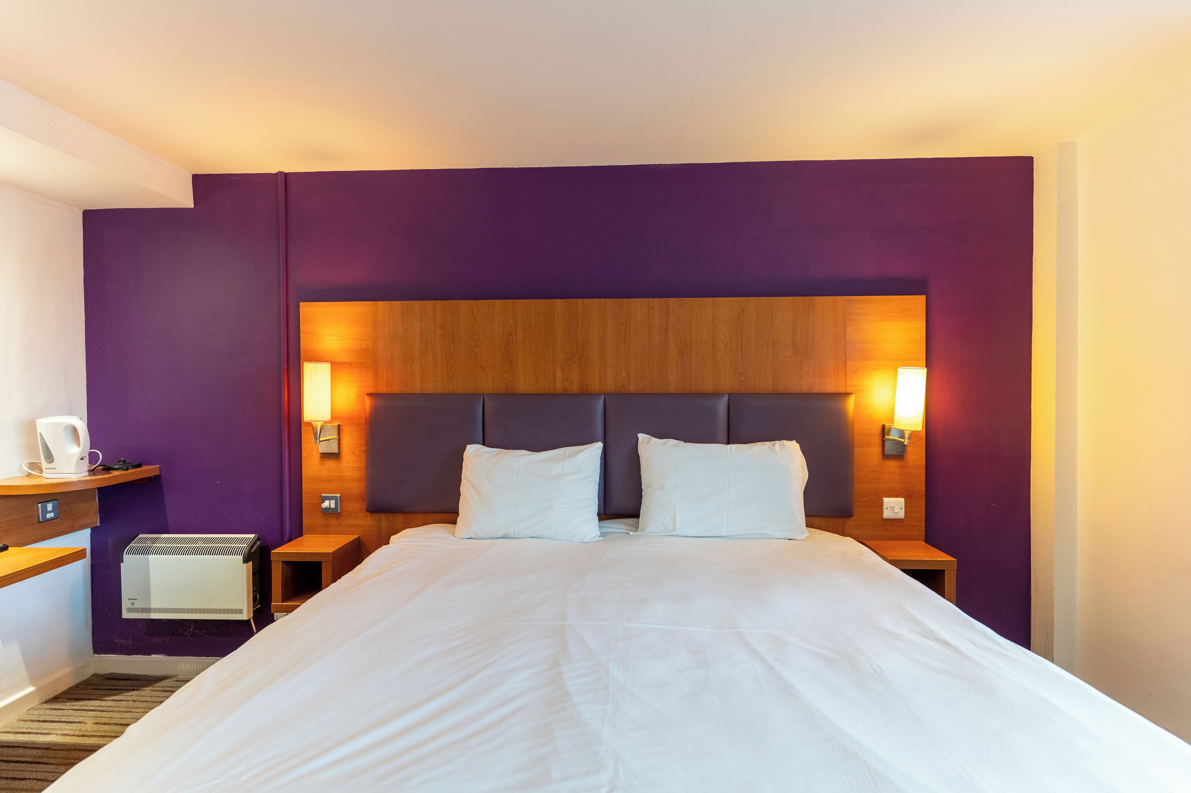 A hotel room with a large bed, purple walls, wooden headboard, bedside lamps, and a small table with a coffee maker.