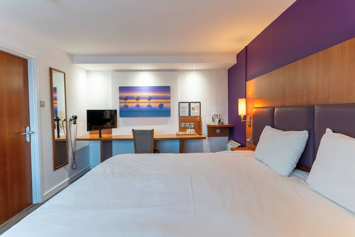 A hotel room with a large bed, a desk with a chair, a television, and a painting on the wall.