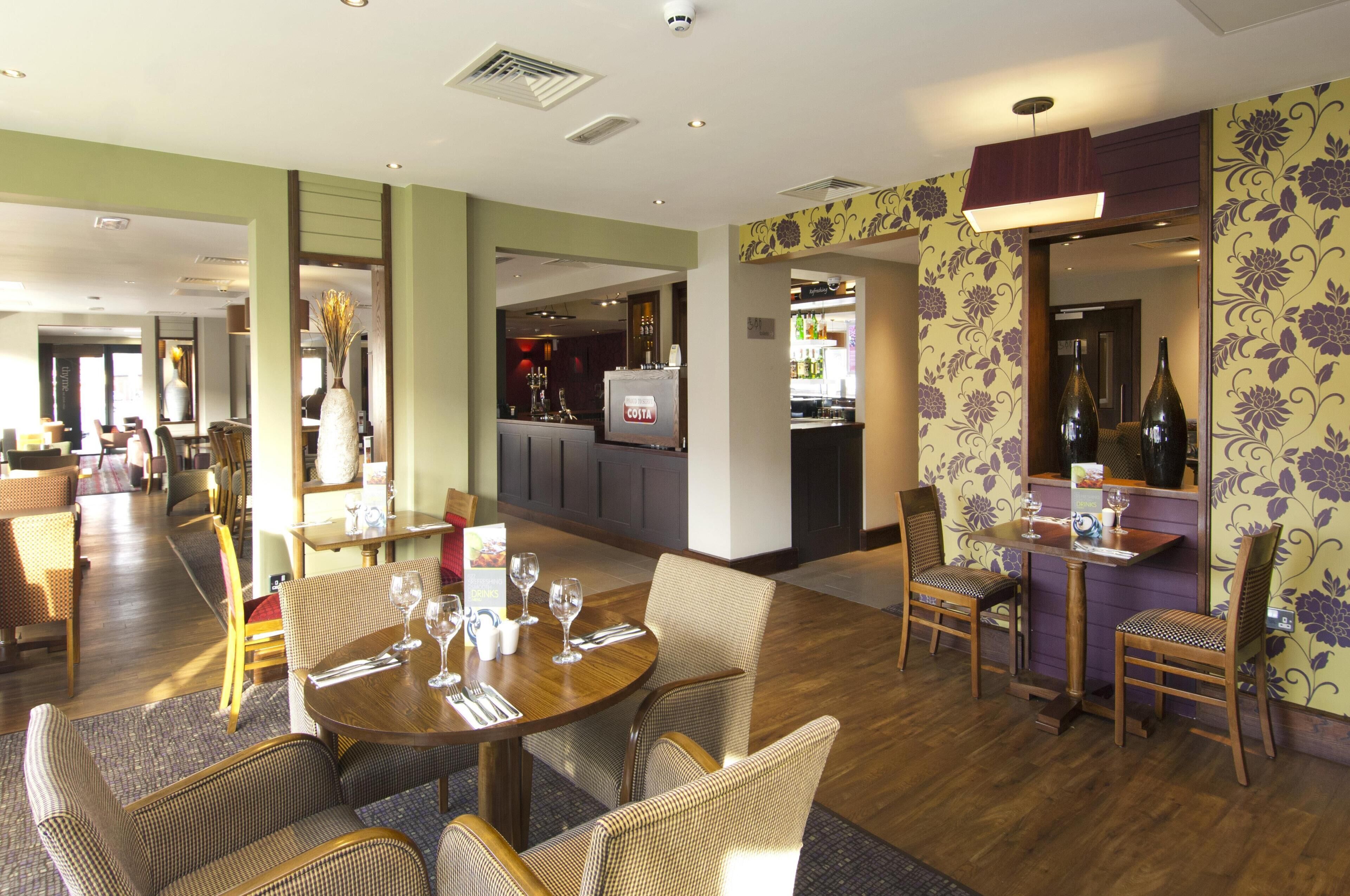 A hotel dining area with round tables, wicker chairs, and a bar area in the background.