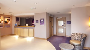 A hotel reception area with a reception desk, a waiting area with a patterned armchair and ottoman, and a door leading to a room. - Premier Inn Camborne (Camborne)