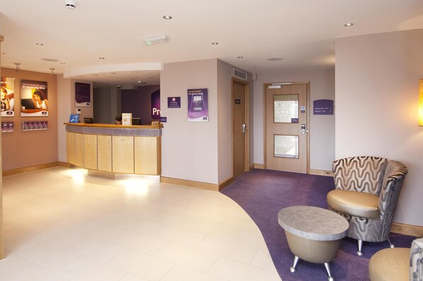A hotel reception area with a reception desk, a waiting area with a patterned armchair and ottoman, and a door leading to a room. - Premier Inn Camborne (Camborne)