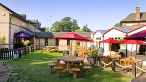 A patio area with wooden tables and red umbrellas, surrounded by brick buildings and a grassy lawn. - Premier Inn Northampton South - Wootton (Northampton)