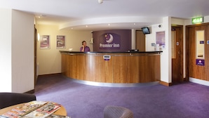 A hotel reception desk with a 'Premier Inn' sign, a reception area with a purple carpet, and a seating area with magazines. - Premier Inn Silverstone (Brackley)