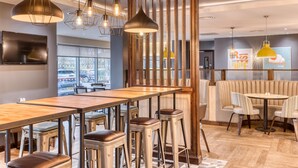 L’intérieur d’un restaurant moderne présente un long bar en bois, des tabourets hauts, un éclairage suspendu et un espace salon avec des tables rondes et des chaises rembourrées.