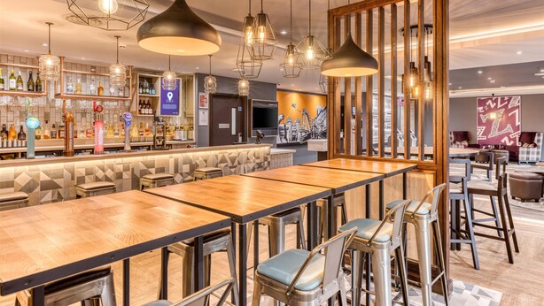 Un espace bar offrant différentes options de sièges, notamment des tabourets de bar et des tables hautes. On y trouve des luminaires suspendus, un comptoir de bar avec diverses bouteilles et un coin salon avec des bancs rembourrés.