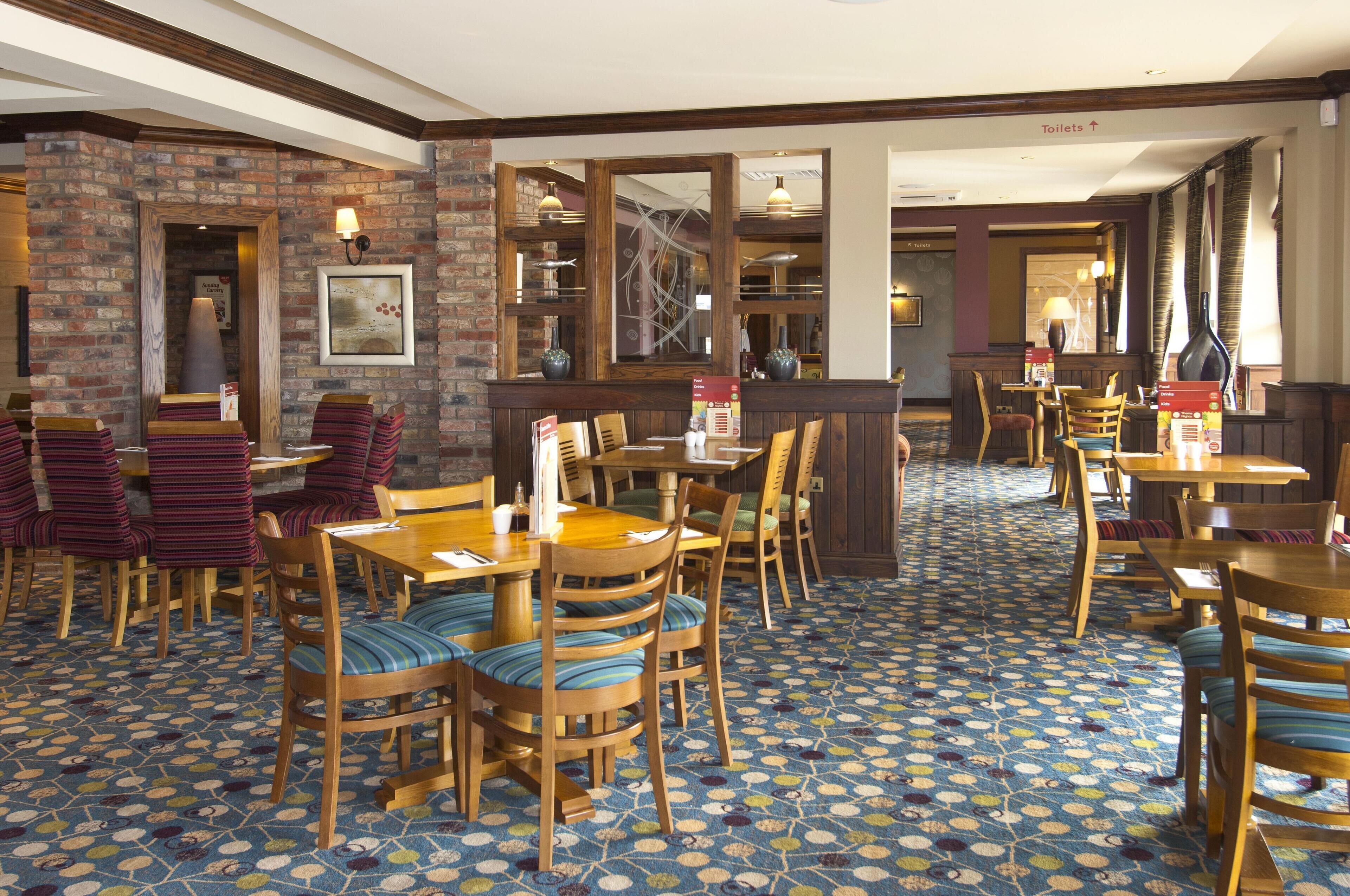 Un restaurant avec des tables et des chaises en bois, un sol à motifs, des murs en briques et un panneau indiquant la direction des toilettes.