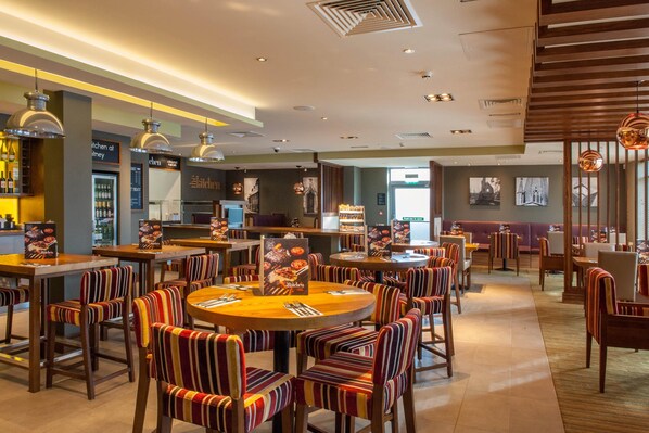 A restaurant with round tables and striped chairs, a bar area, and a view of the city. - Premier Inn Witney (Witney)
