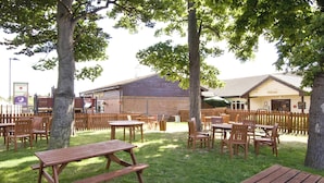 An outdoor seating area with wooden tables and chairs, a brick building with a 'Welcome' sign, and a fenced area. - Premier Inn Sittingbourne Kent (Sittingbourne)