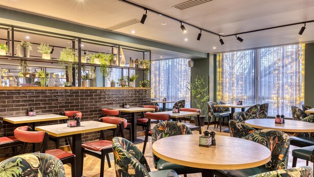 A restaurant with round tables and chairs, a brick wall, and shelves with plants. - Premier Inn Margate (Margate)