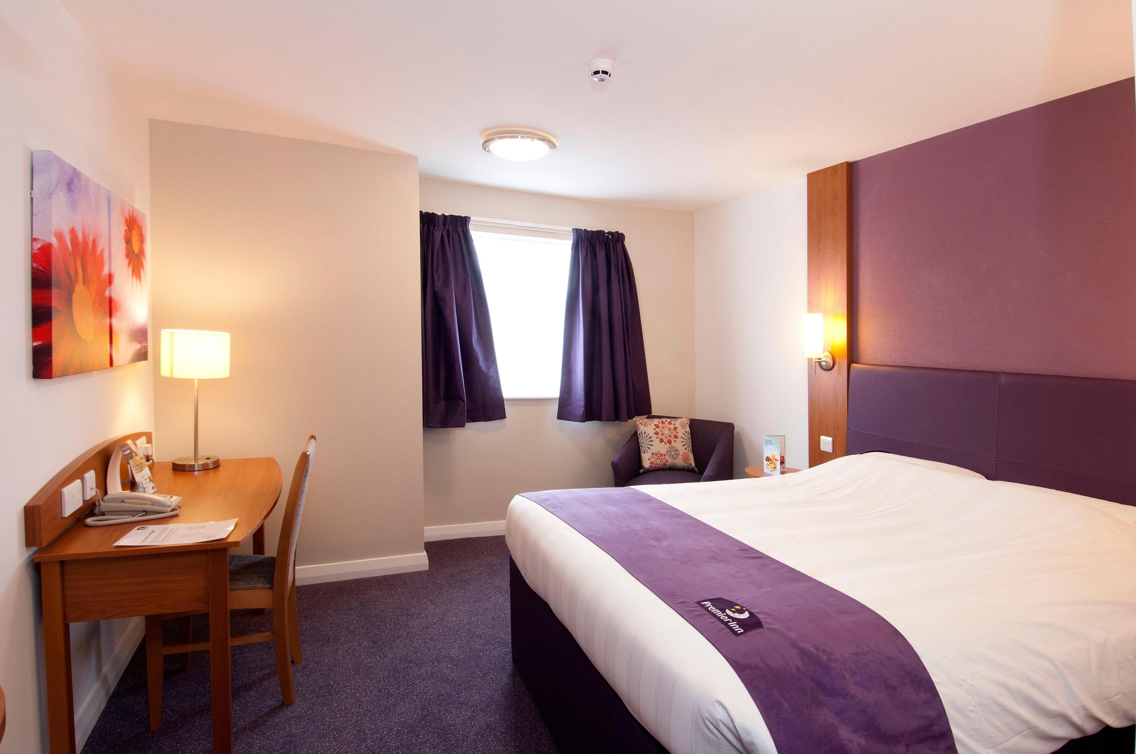 A hotel room with a bed, a desk, a chair, a window with curtains, a wall-mounted lamp, and a ceiling light.
