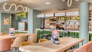 A modern bar area with wooden tables and chairs, a bar counter with various bottles and glasses, and hanging plants.