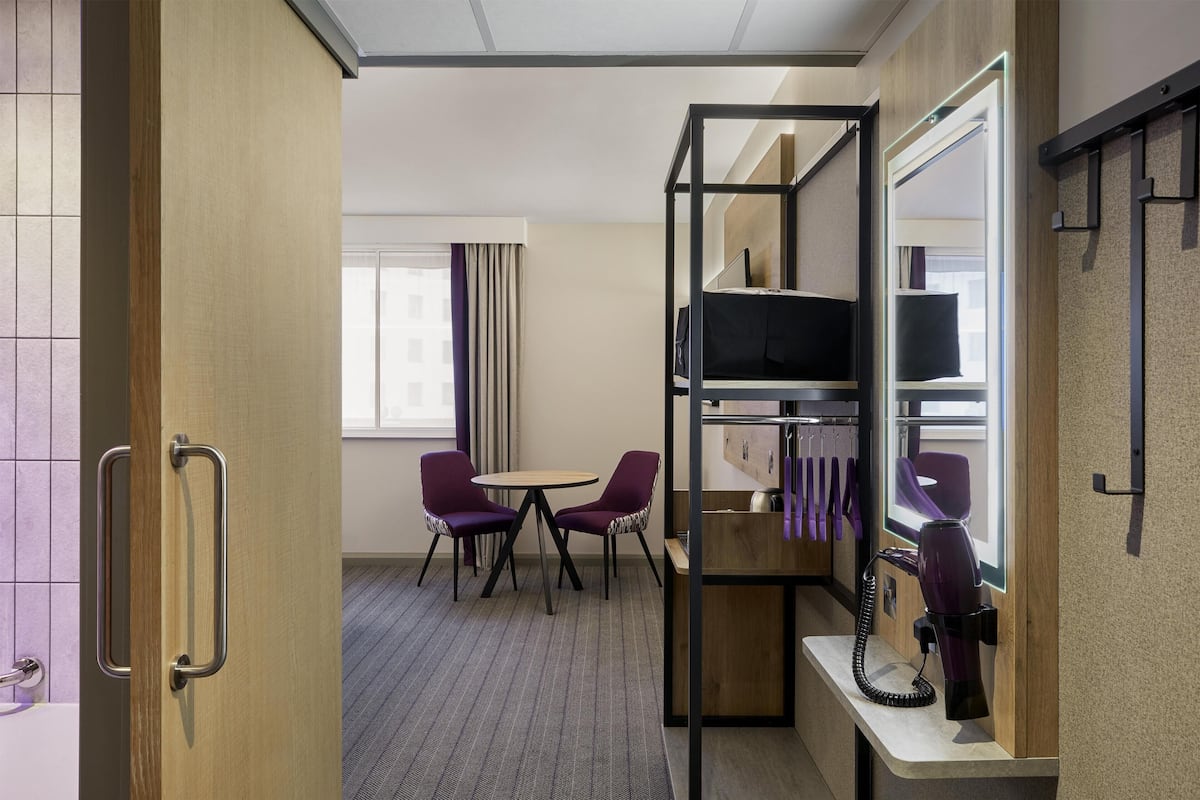 A hotel room with a bed, a TV, a desk with a hairdryer, two purple chairs, and a window with curtains.