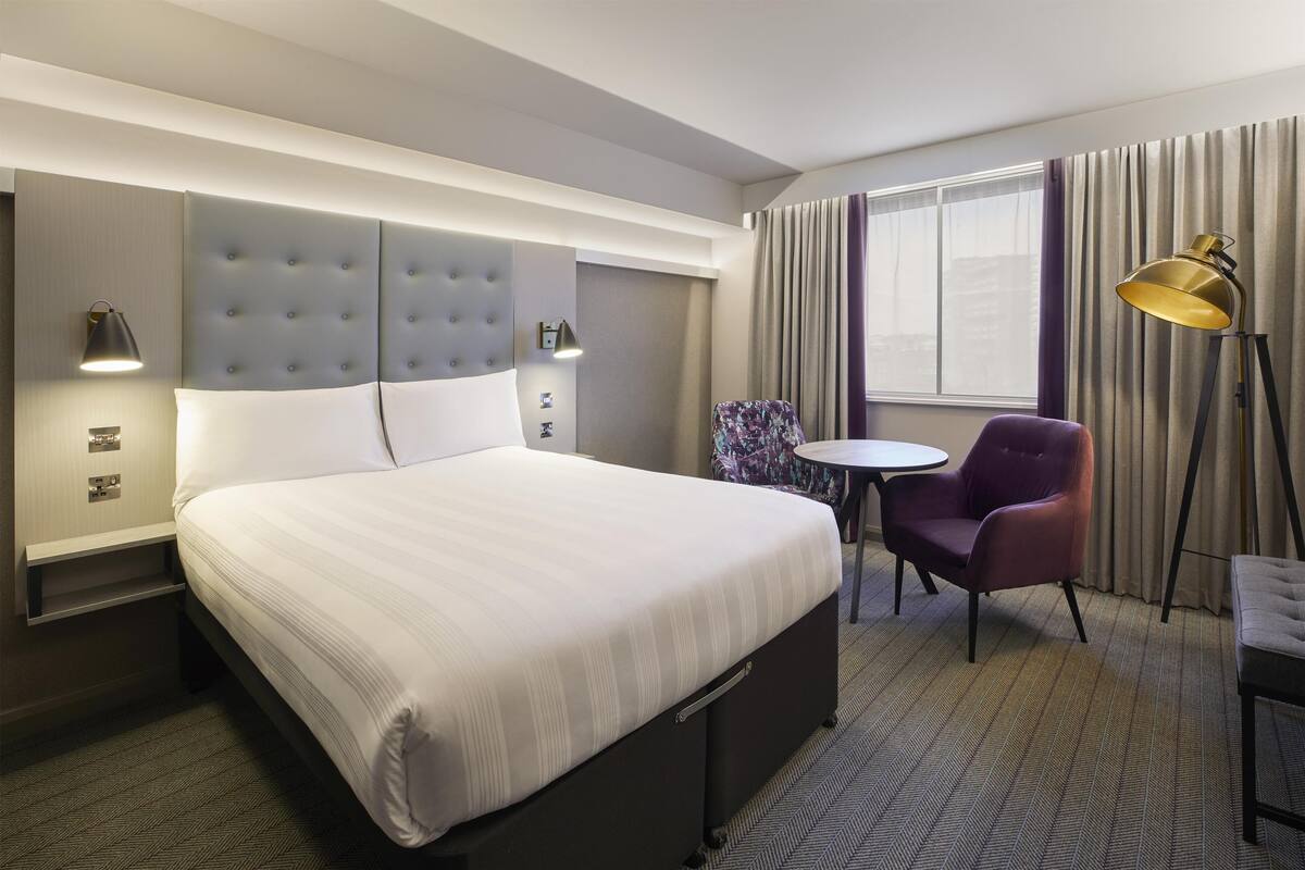 A hotel room with a large bed, bedside lamps, a small table, a chair, and a floor lamp.