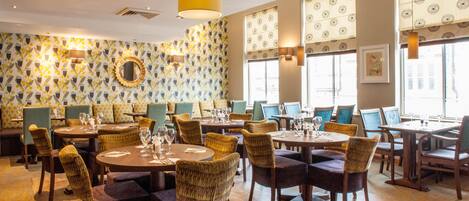 A restaurant with round tables, wicker chairs, patterned wallpaper, and large windows.