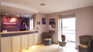 A hotel reception area with a counter, a receptionist, two patterned armchairs, and a window with a view. - Premier Inn Llanelli Central West (Llanelli)