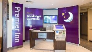 Ein Empfangsbereich von Premier Inn mit einem 'Check In'-Schalter, einem digitalen Bildschirm und einem Schild mit der Aufschrift 'Good Night Guarantee'.