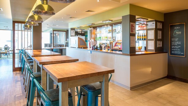 A restaurant interior with multiple wooden tables and blue bar stools, a bar area with various bottles, and a menu board on the wall. - Premier Inn Winchester (Winchester)