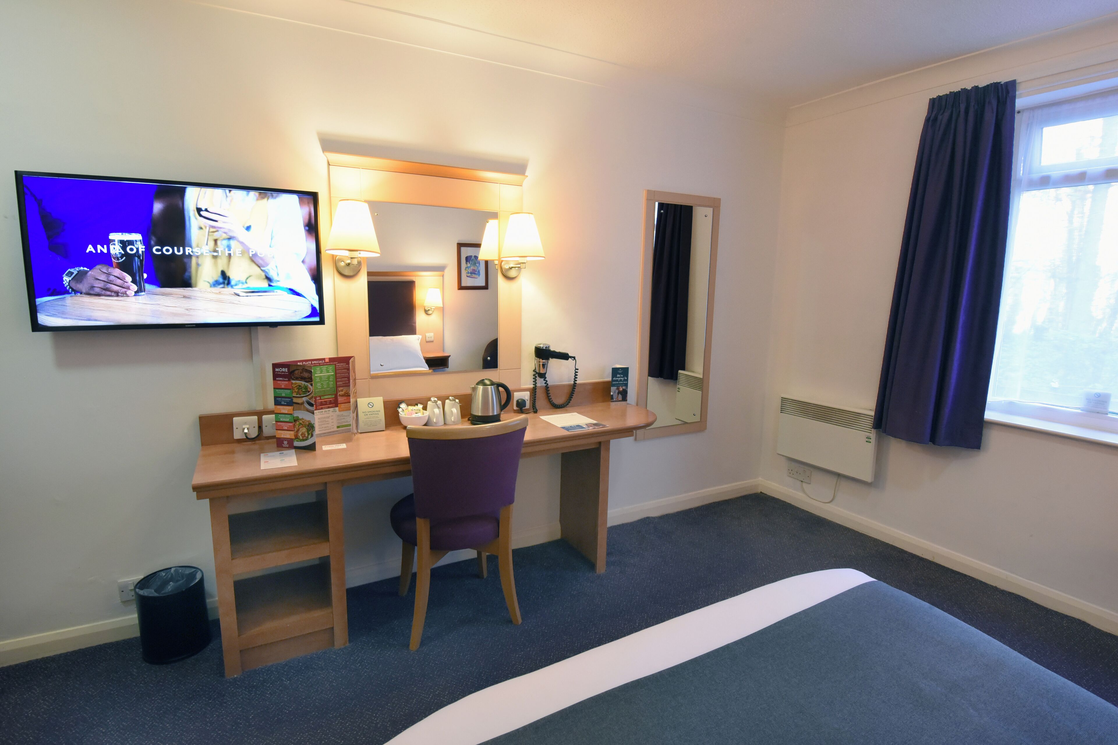 Habitación de hotel con escritorio, silla, televisión, espejo y ventana con cortinas.