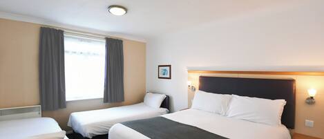A hotel room with two single beds, a large bed, a window with curtains, a picture on the wall, and a nightstand with a lamp.