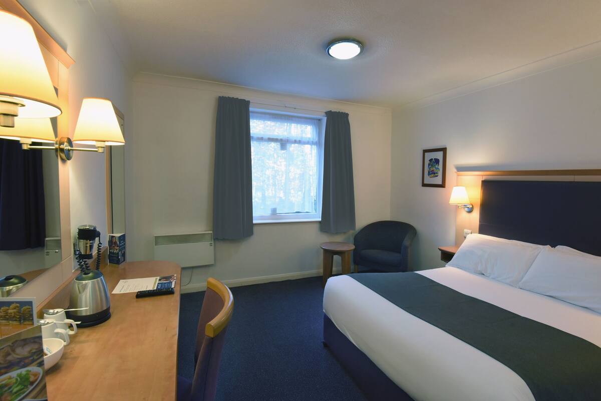 A hotel room with a bed, a desk with a kettle, a chair, a small table, a window with curtains, and a picture on the wall.