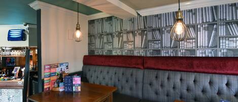A cozy bar area with a wooden counter, a cushioned bench, and a wall with bookshelves.
