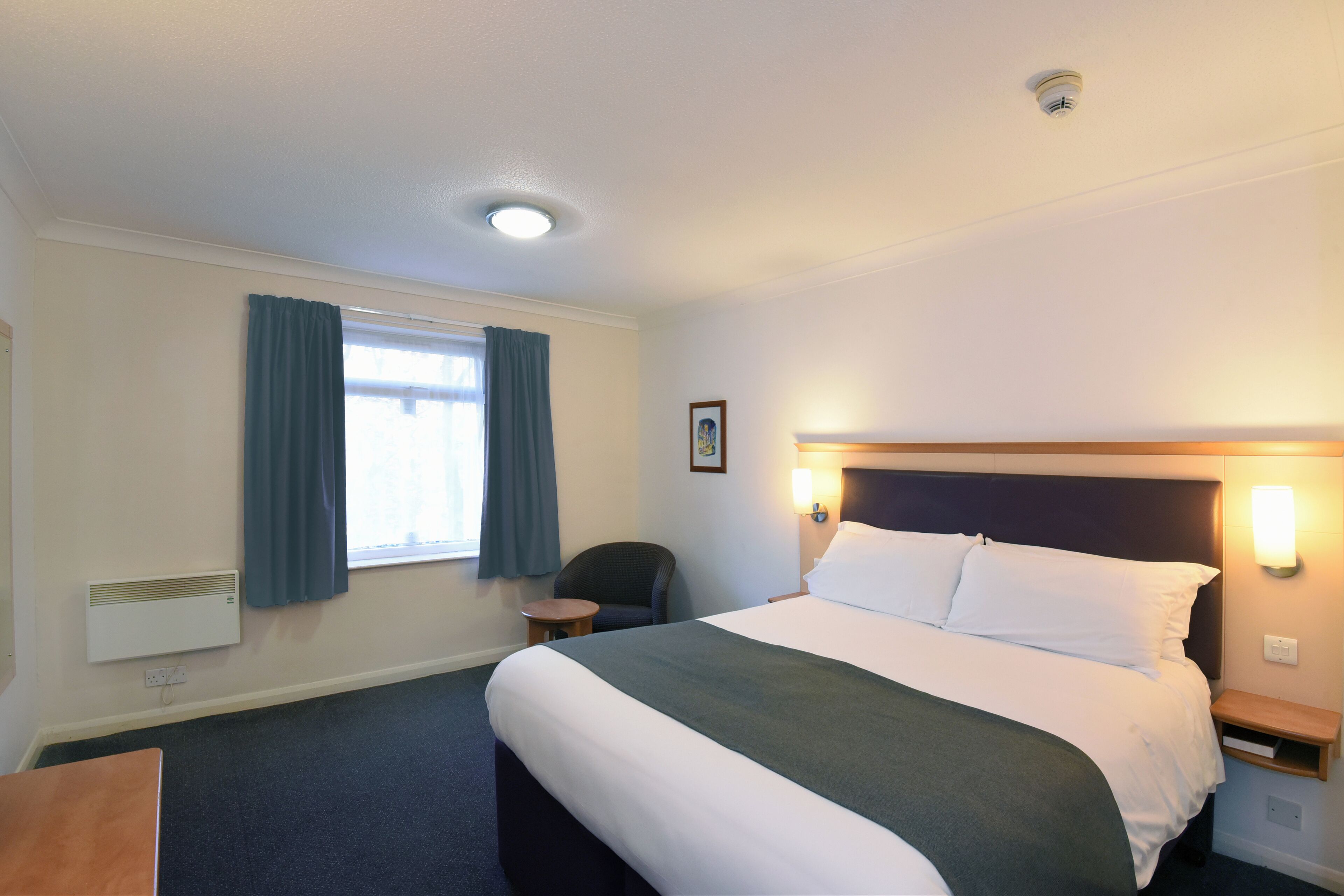 A hotel room with a large bed, a bedside table, a chair, a window with curtains, and a wall-mounted heater.