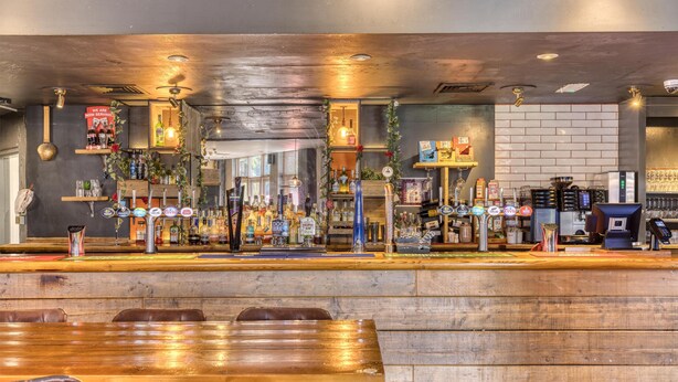 A bar area with wooden furniture, a counter with various bottles, and a coffee machine. - Premier Inn Basingstoke West (Basingstoke)