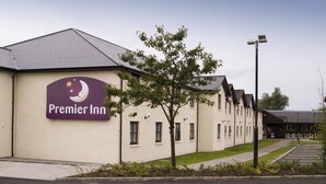 A hotel with a sign reading 'Premier Inn,' a parking lot, and a building with a sloped roof. - Premier Inn Glasgow - Motherwell (Motherwell)