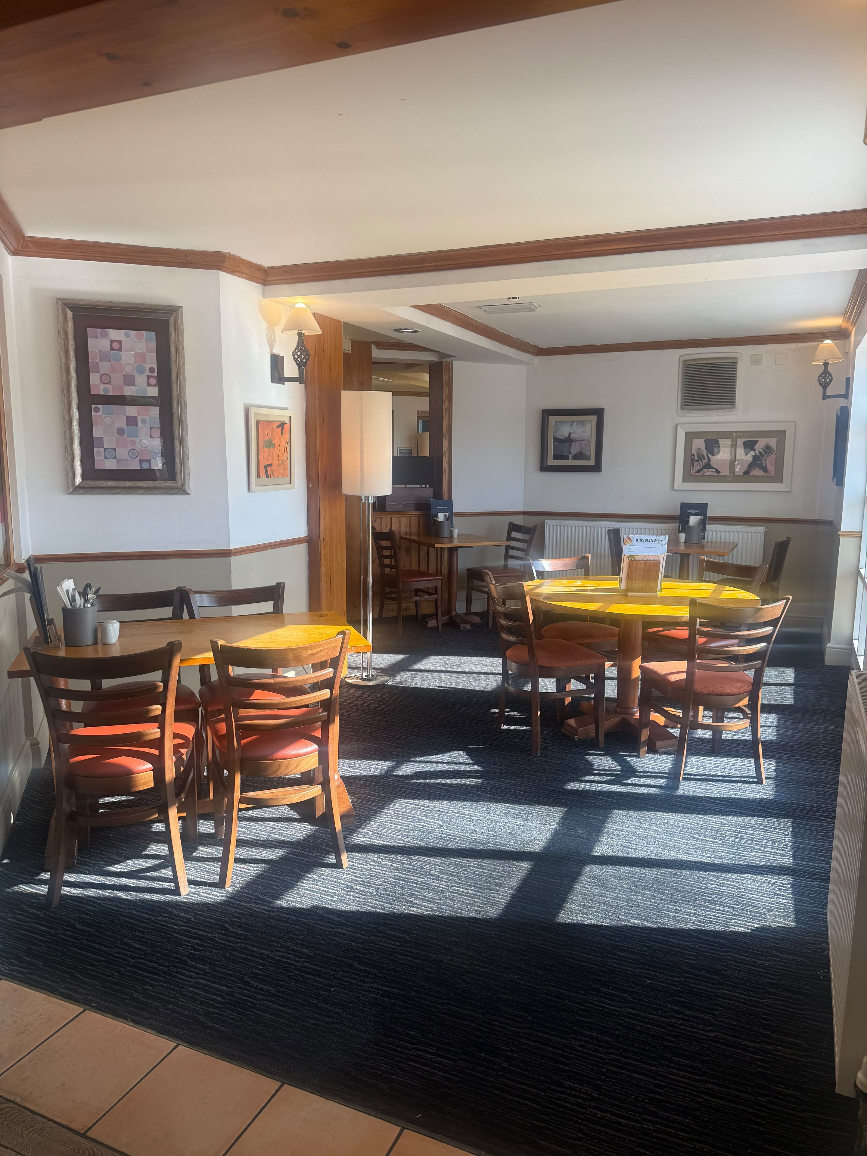 Uma área de refeições com mesas e cadeiras de madeira, uma máquina de café e quadros nas paredes.