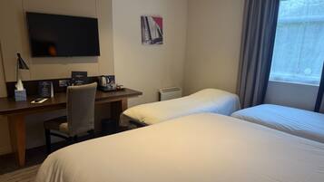 A hotel room with a desk, two beds, a TV, and a window with curtains.