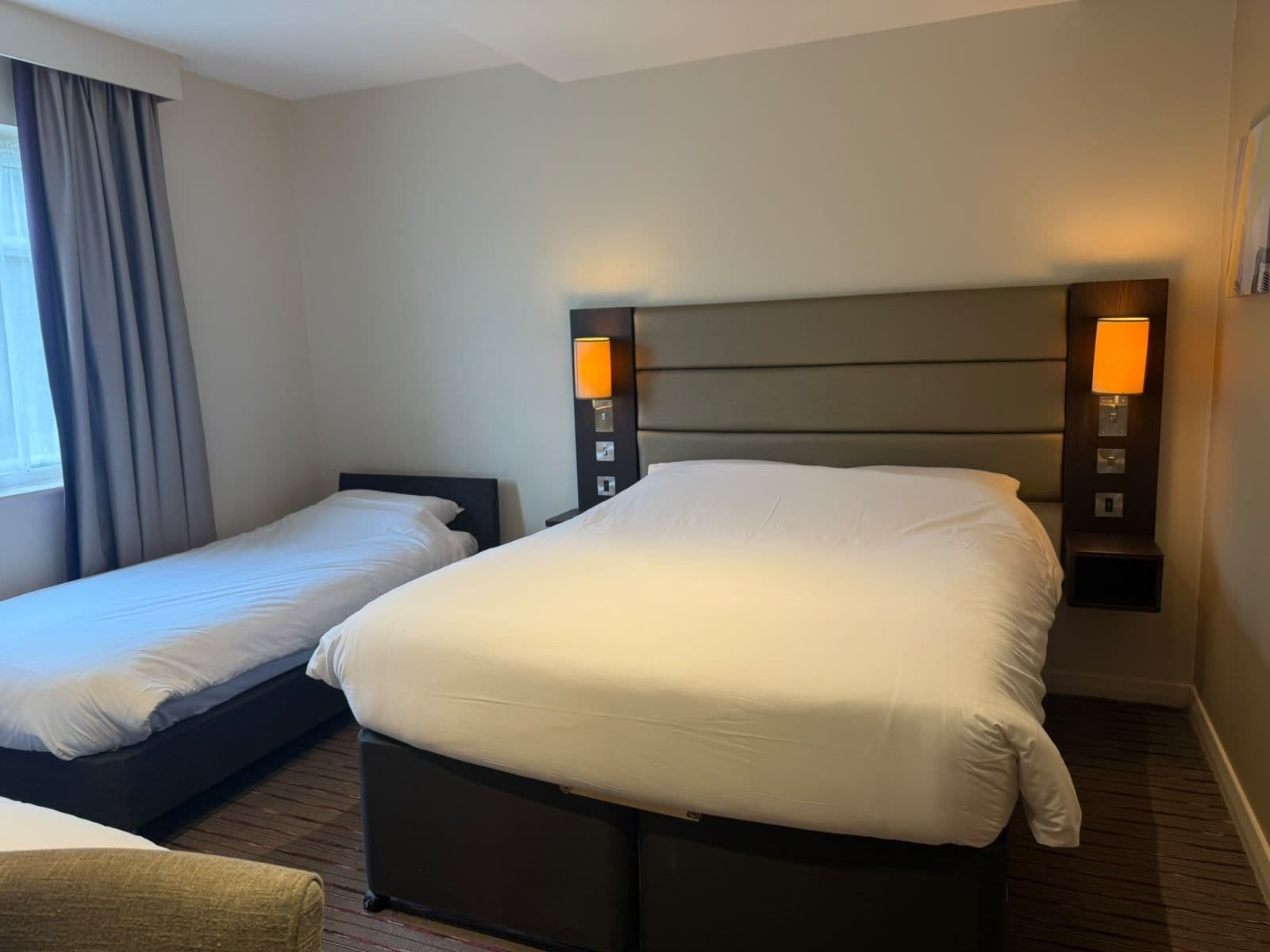 A hotel room with two beds, a nightstand with a lamp, and a window with curtains.