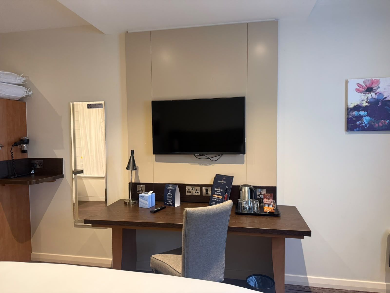 Quarto de hotel com uma cama, escrivaninha com cadeira, televisão na parede, cafeteira e um quadro na parede.