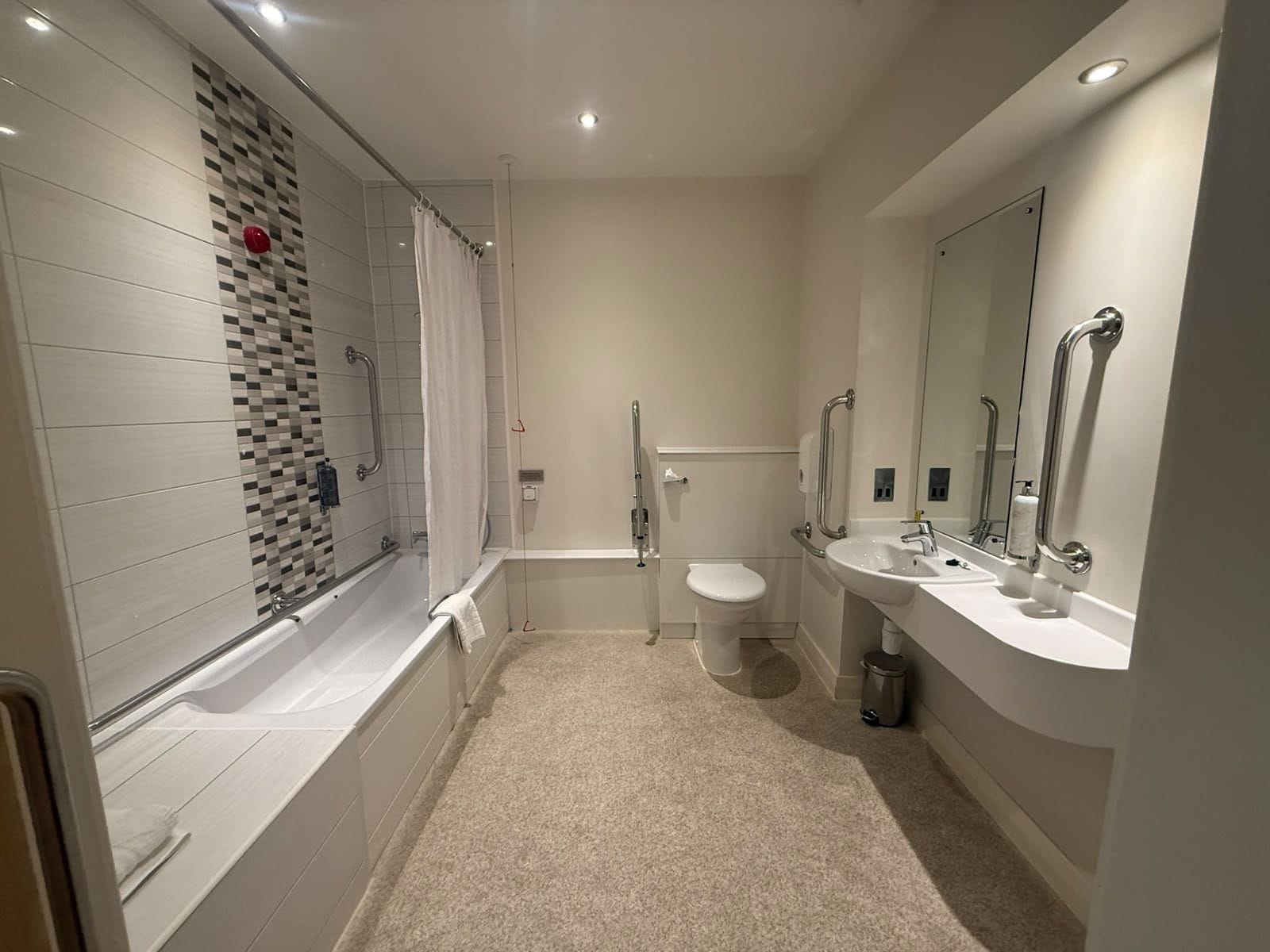 Ein modernes Badezimmer mit Badewanne, Toilette und Waschbecken, ausgestattet mit Haltegriffen und einer Duschvorhang.